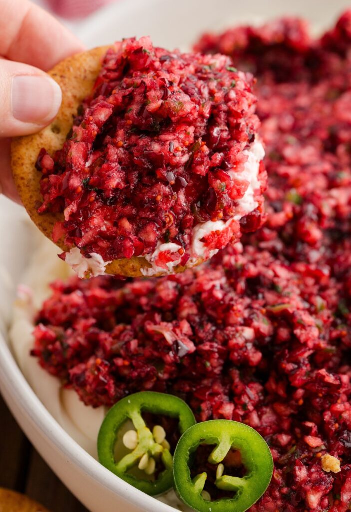 Cream cheese dip with a cranberry and jalapeno topping, a hand holding a cracker ready to dip.