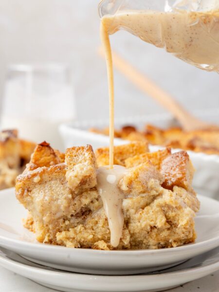 A serving of the bread pudding a white plate with the glaze being poured over top of it from a glass.