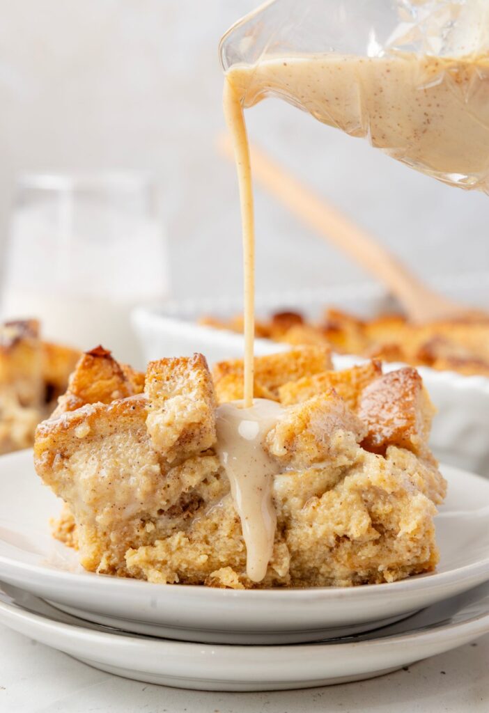 A serving of the bread pudding a white plate with the glaze being poured over top of it from a glass. 