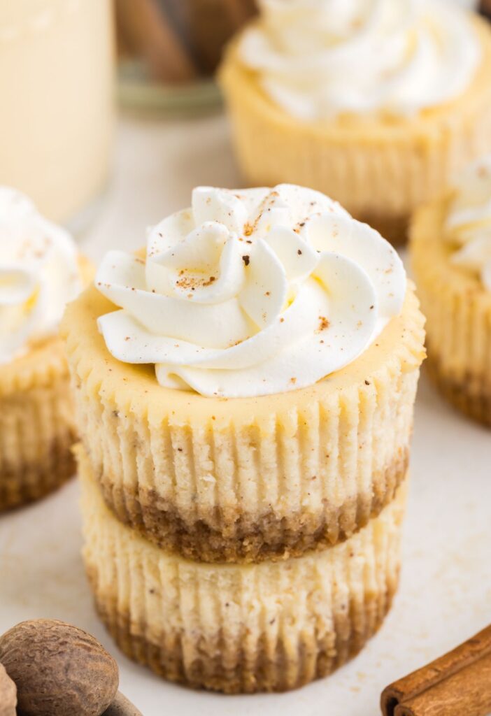 Baked mini eggnog cheesecakes with whipped cream on top and cinnamon sticks beside the plate of mini cheesecakes.