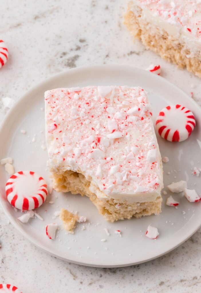 White plate of a Krispie treat with a peppermint candy next to it, and a bite taken out of it to show the gooey center. 
