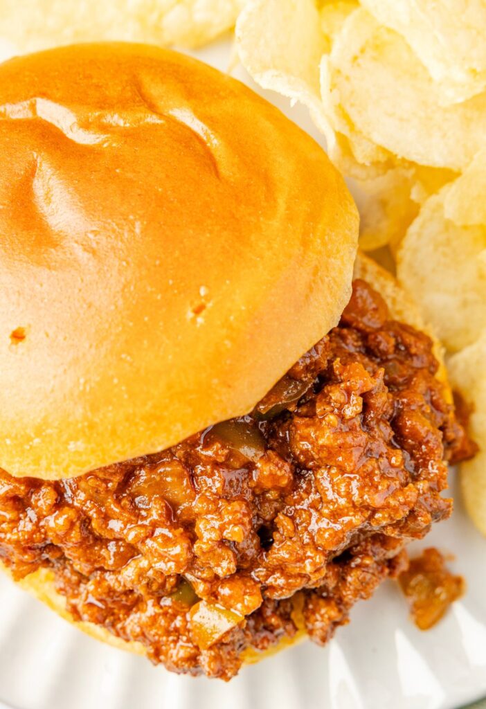 A sloppy Joe sandwich in a soft bun with potato chips on the side of the plate.