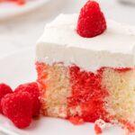 A slice of poke cake with a raspberry on top of whipped cream frosting.