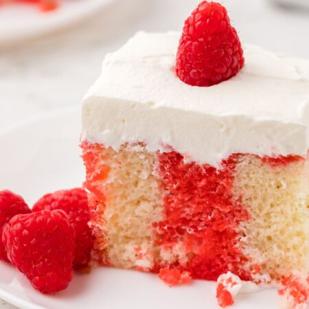 A slice of poke cake with a raspberry on top of whipped cream frosting.