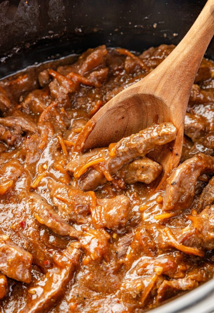 Inside of the slow cooker with the cooked beef in a wooden serving spoon.