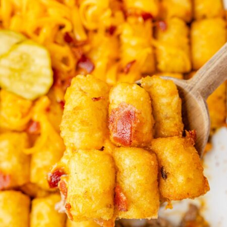 A casserole pan of the tater tot casserole with a wooden serving spoon with a scoop on it.