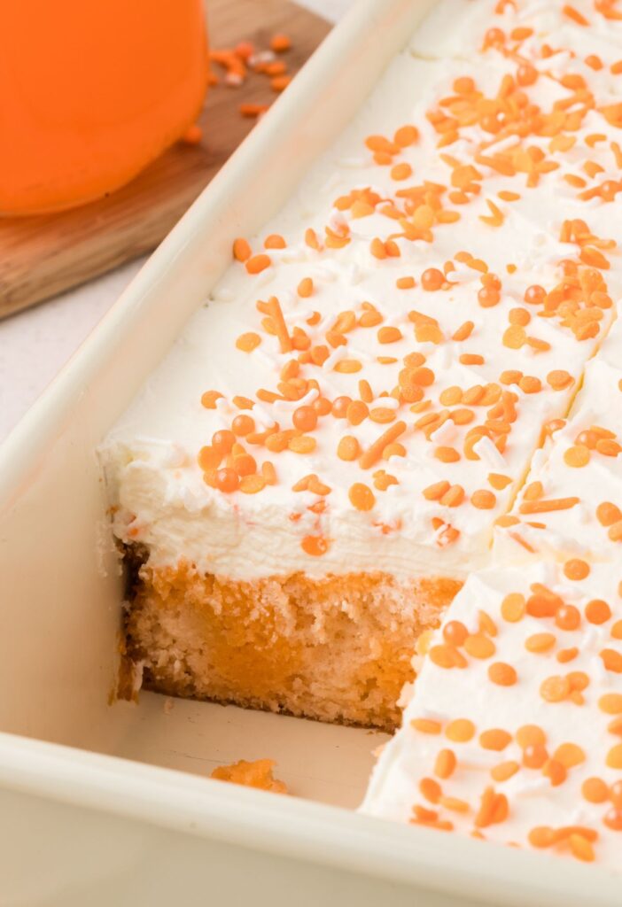 A cake pan with the baked poke cake inside of it, topped with whipped cream and orange sprinkles.