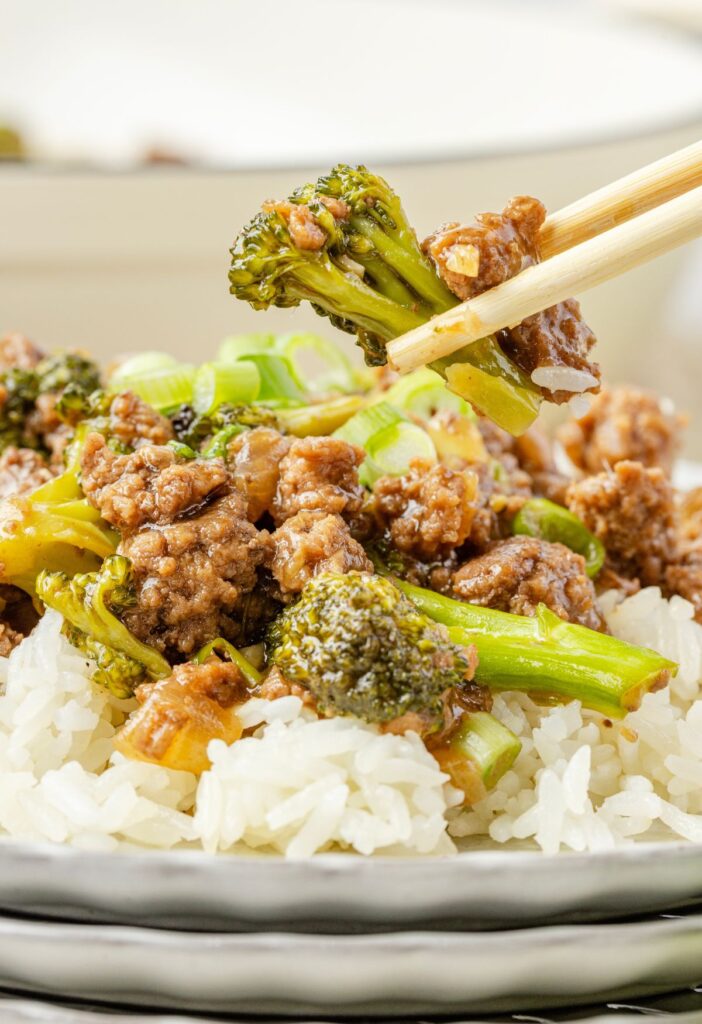 Serving of ground beef and broccoli on white rice with chopsticks holding a bite