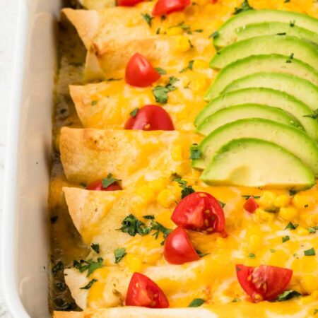 A white pan of chicken enchiladas topped with cheese, avocado, and corn.