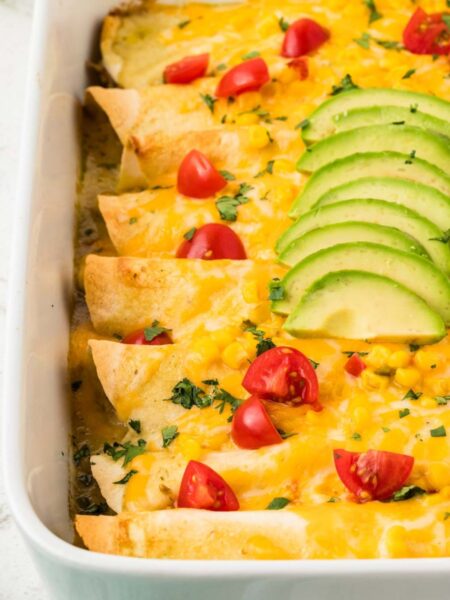 A white pan of chicken enchiladas topped with cheese, avocado, and corn.