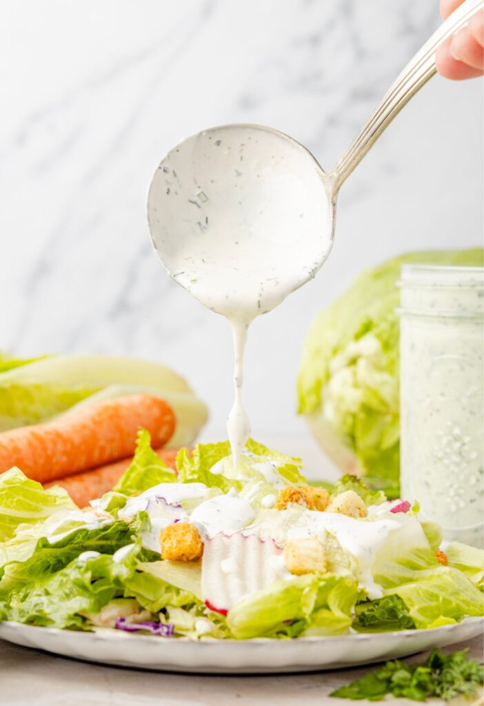 A salad on a plate with a spoon above it, with ranch on it, and the ranch is being poured over top the salad.