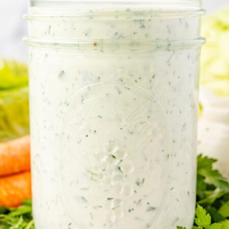 A clear jar of the ranch dressing in it with some fresh herbs in the background.