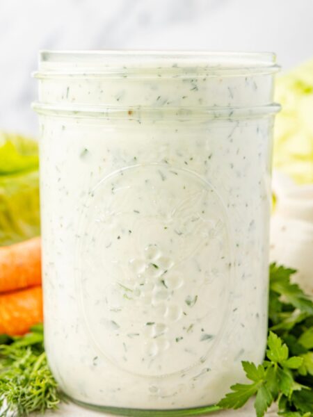 A clear jar of the ranch dressing in it with some fresh herbs in the background.