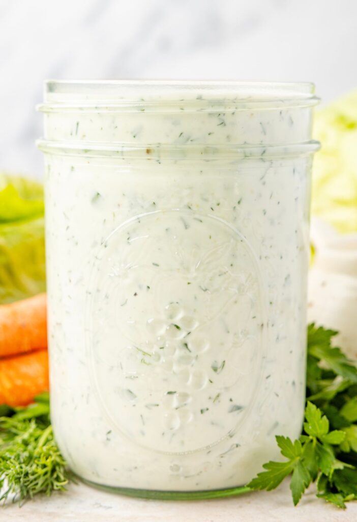 A clear jar of the ranch dressing in it with some fresh herbs in the background.