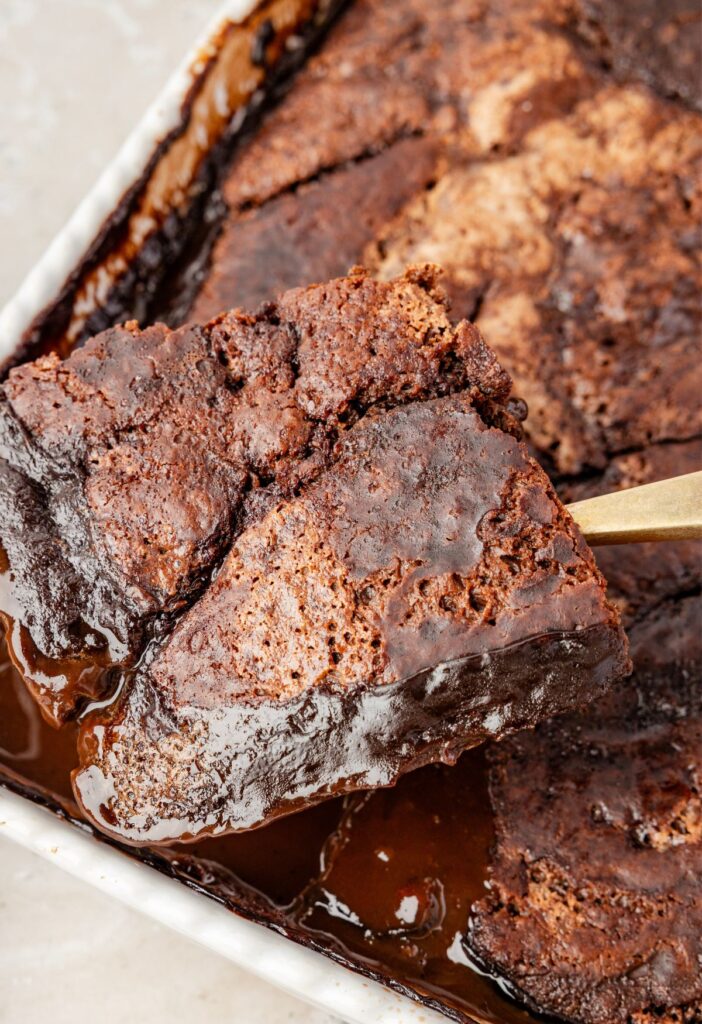 A white pan with the chocolate cobbler inside of it, the hot fudge sauce, and a serving spoon taking a scoop of it.