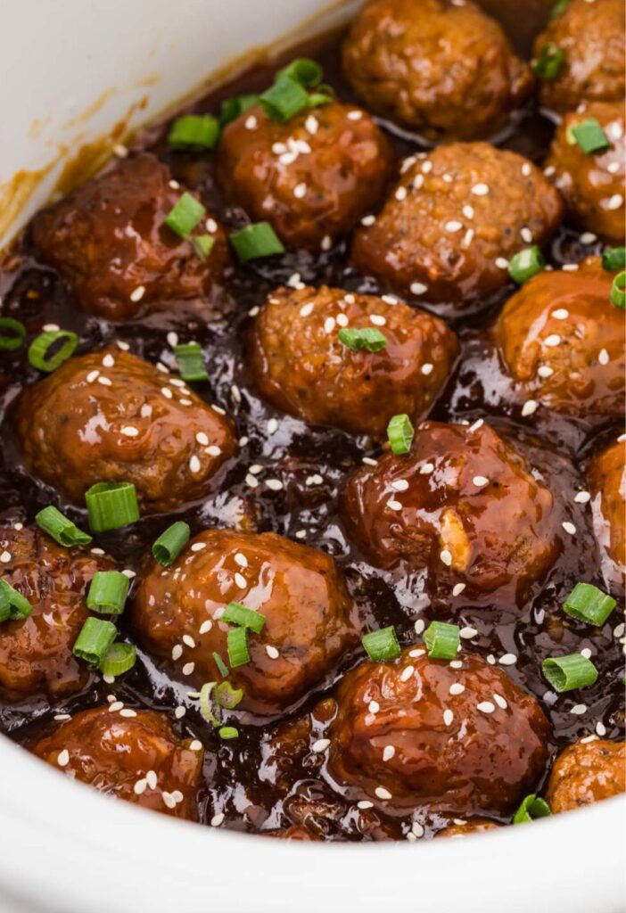 A crockpot full of cooked orange chicken meatballs with green onions and sesame seeds on top