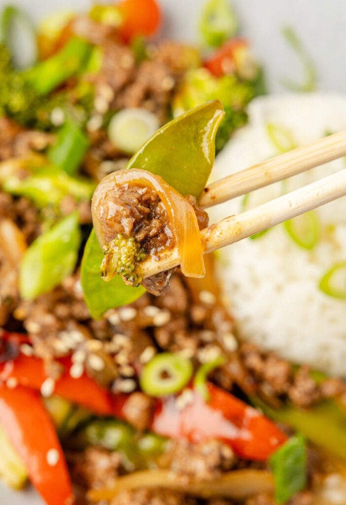 Chopsticks picking up some stir fry above the plate of stir fry made with ground beef and white rice for serving.