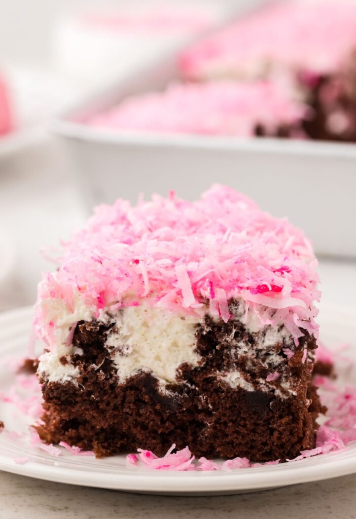 A slice of the poke cake that looks like a hostess snoballs poke cake sitting on a plate with the cake pan behind it in the background.