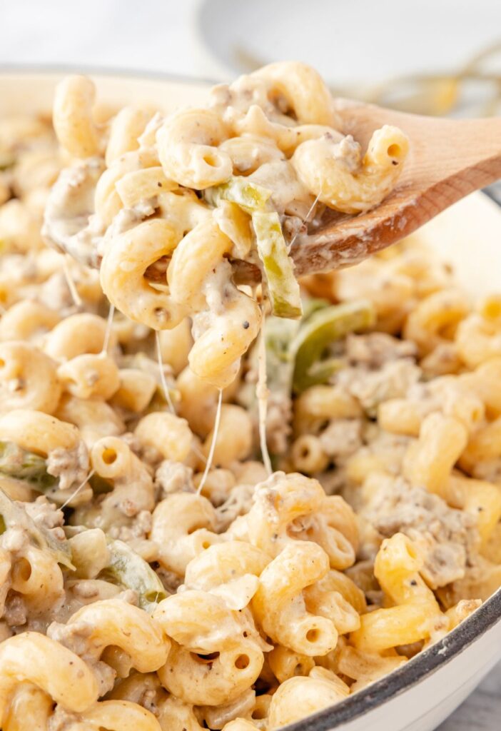 A skillet of cheesesteak pasta with a wooden spoon and a cheese pull with some pasta on it