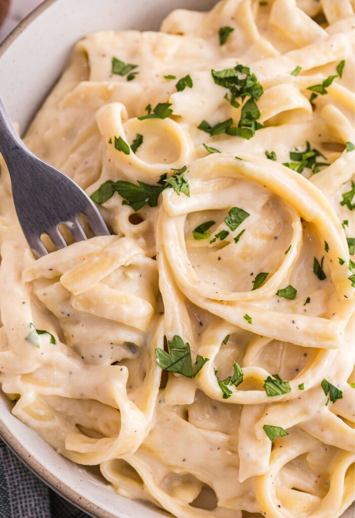 A bowl of fettuccine Alfredo sauce with a fork and fresh parsley on top.