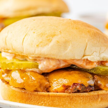 A burger with cheese and pickles and sauce, on a hamburger bun sitting on a plate with fries.