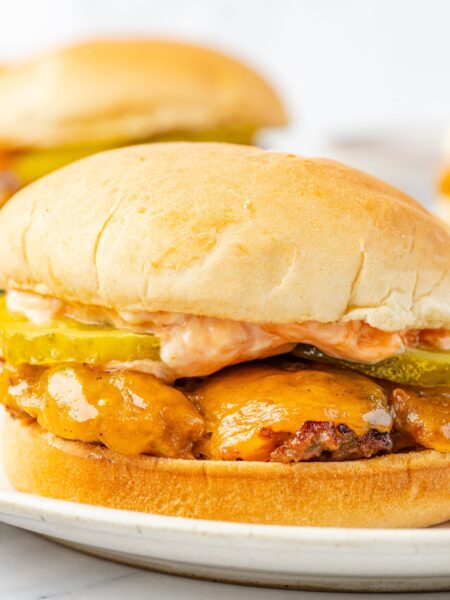 A burger with cheese and pickles and sauce, on a hamburger bun sitting on a plate with fries.