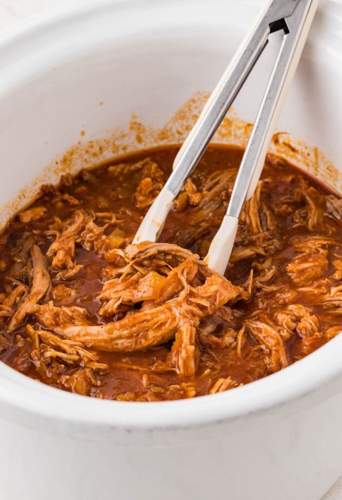 A crockpot insert with shredded pork and tongs for serving.