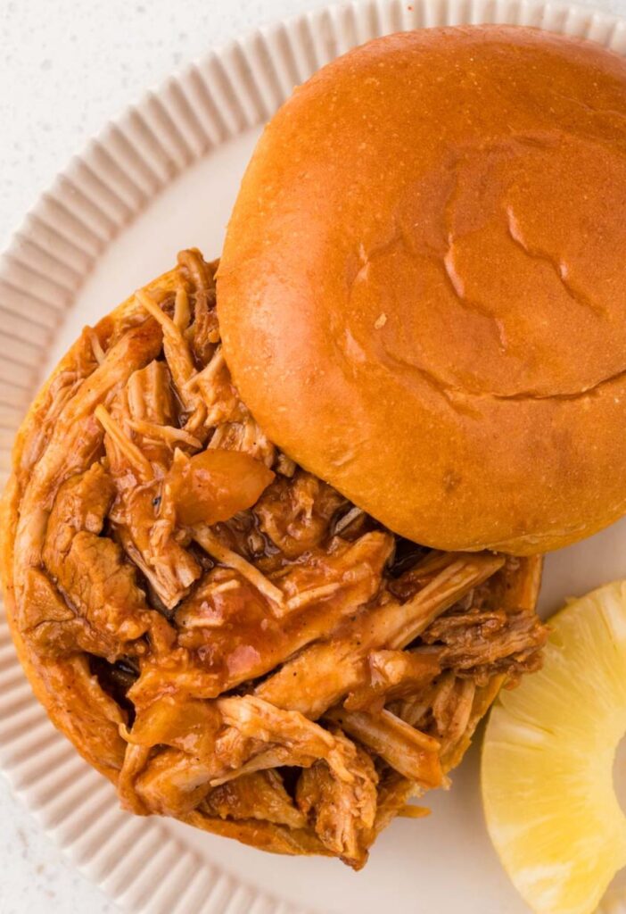 Open faced pulled pork sandwich with pineapple next to it on a white plate.