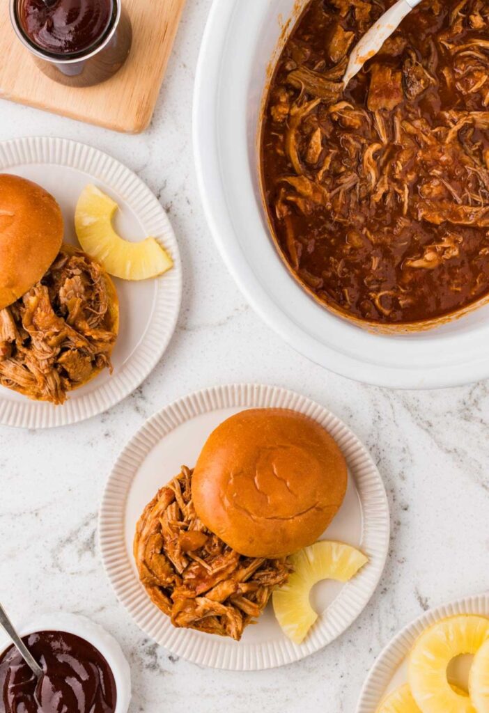 Top view of the crockpot of pulled pork and a serving of it on a brioche bun.