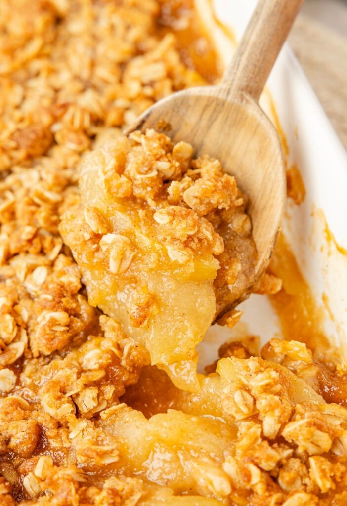 A wooden spoon holding some of the applesauce crisp on it above the pan of apple crisp below it.