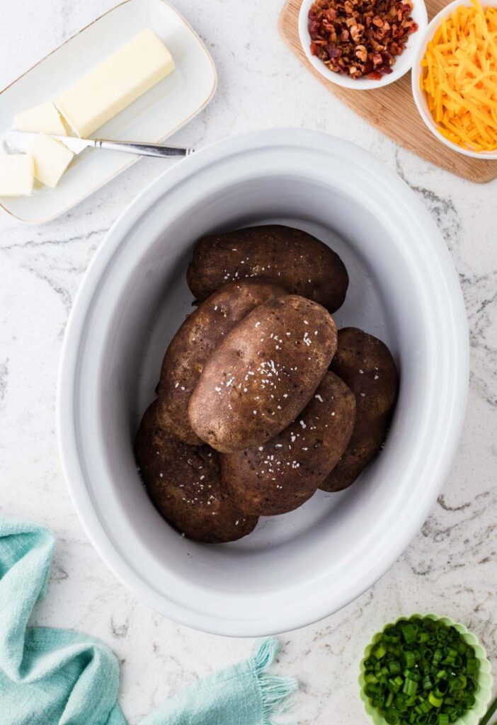 A slow cooker with baked potatoes inside and toppings to the sides of the slow cooker.