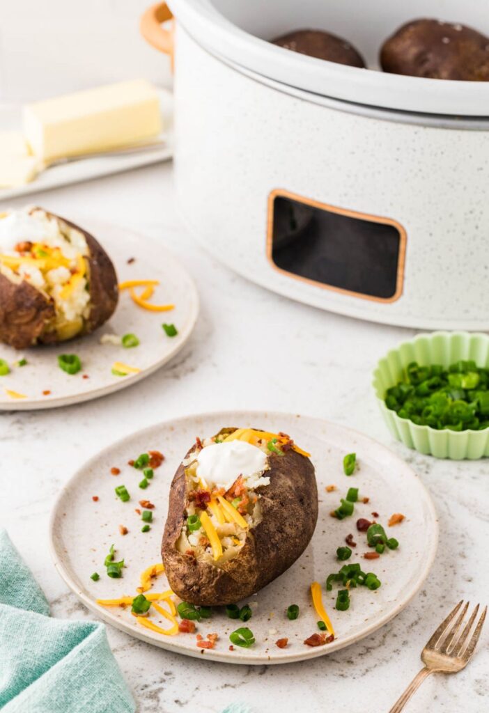 A slow cooker in the background with a baked potato on a plate out in front.