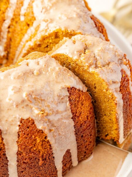 Baked pumpkin bundt cake on a white plate with a glaze, a serving spatula taking a slice from the cake.