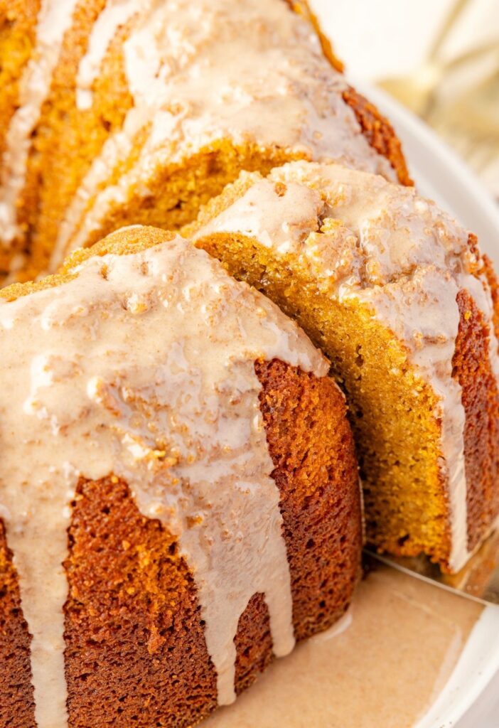 Baked pumpkin bundt cake on a white plate with a glaze, a serving spatula taking a slice from the cake.
