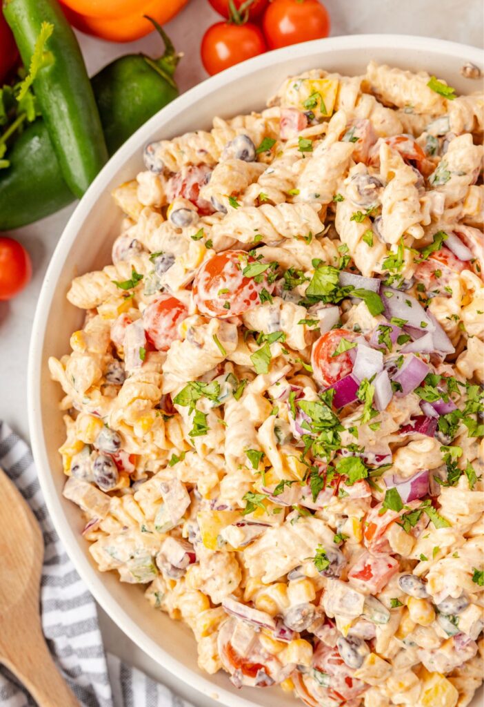 A white serving bowl of pasta salad with fresh vegetables above the bowl for decoration.