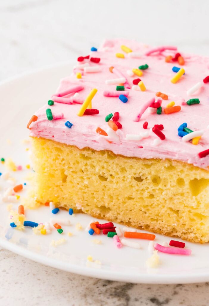 A slice of sugar cookie cake on a white plate with pink frosting and sprinkles.