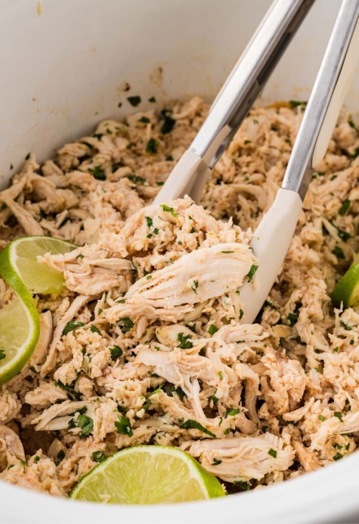 Shredded cilantro lime chicken inside the crockpot with tongs scooping some up.