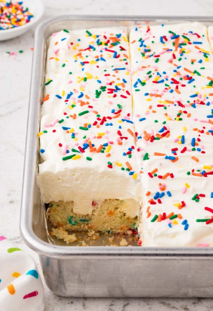 A cake pan with the funfetti cake inside, a piece cut out so you can see the poked hole filling.