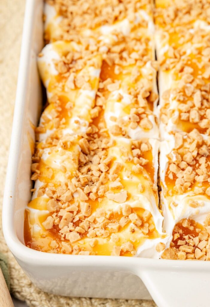 A cake pan with the baked pumpkin poke cake inside of it topped with whipped cream and toffee bits.