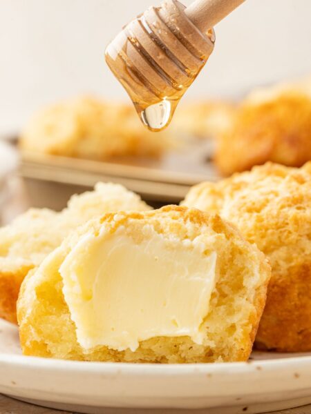 A sour cream biscuit cut in half with butter on it and honey being dropped onto it.