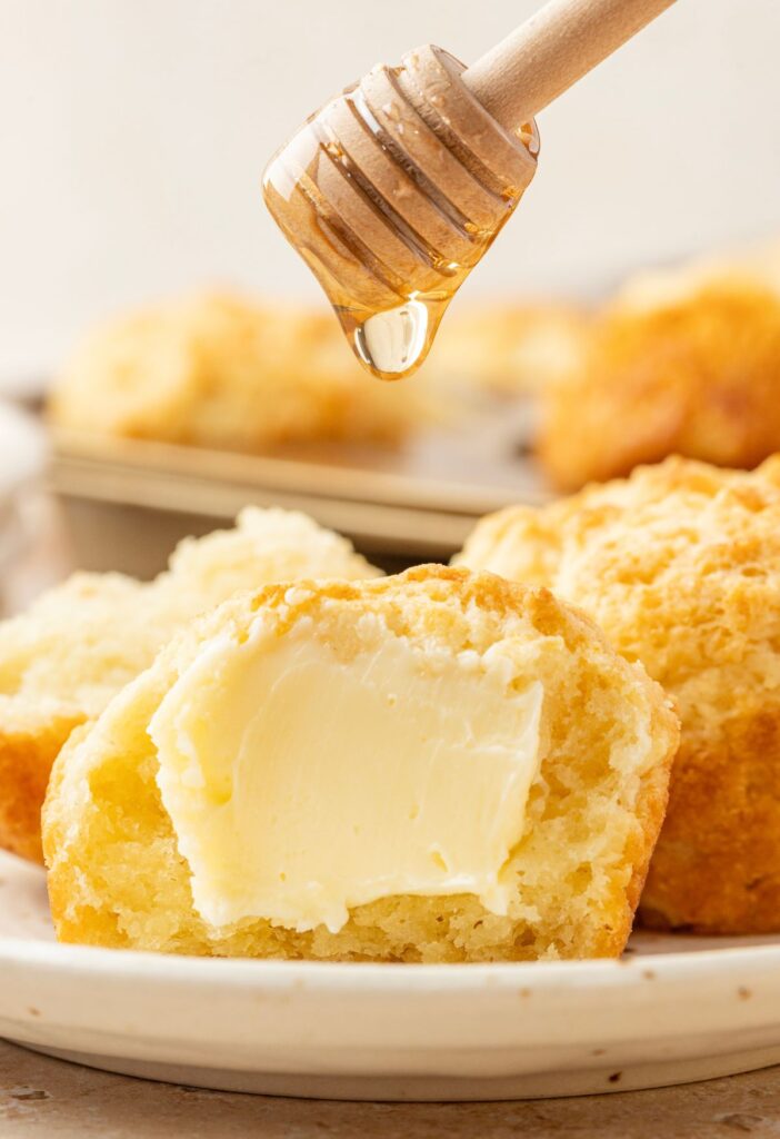 A sour cream biscuit cut in half with butter on it and honey being dropped onto it.