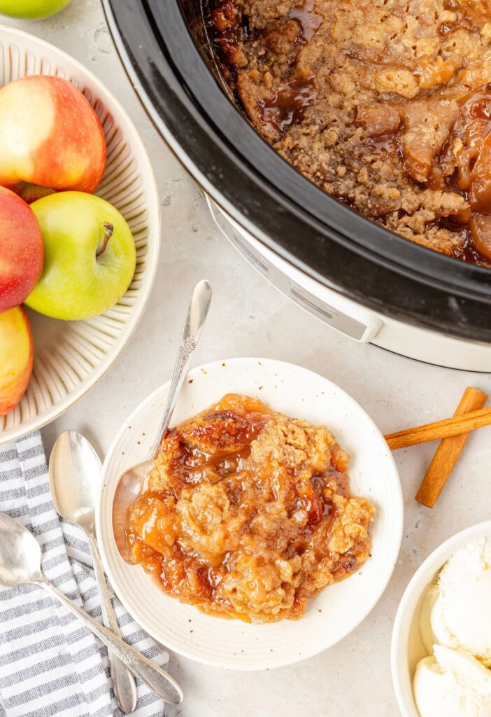 Top view of the slow cooker with the apple crumble inside and fresh apples to the side.